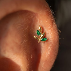 May include: A gold ear piercing stud shaped like a small branch with two marquise-cut green gemstones set as leaves. The jewellery features a polished gold metal finish and is positioned on the cartilage of an ear, showcasing a botanical design.