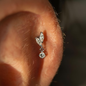 Peut inclure: Boucle d'oreille en argent avec un motif en forme de feuille et une gemme ronde pendante. Le bijou est serti de pierres claires et scintillantes. Le bijou est porté sur une oreille.