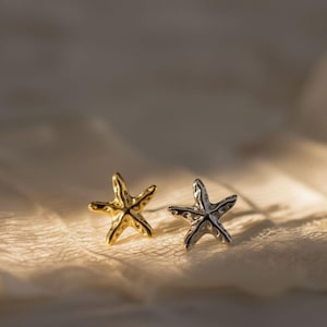 以下が含まれることがあります： ヒトデの形をしたスタッドピアス2点。1点は光沢のあるゴールドトーン、もう1点は光沢のあるシルバートーンで仕上げられています。それぞれのピアスは、ヒトデの自然な形を捉えた、質感のある有機的な表面と5本の腕が特徴です。