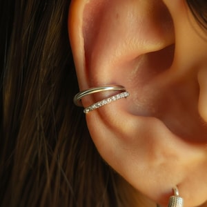 May include: A silver-toned double-band ear cuff featuring one smooth, plain band and one thinner band encrusted with small, clear, sparkling stones. The jewelry is positioned on the cartilage of the ear, creating a layered appearance.