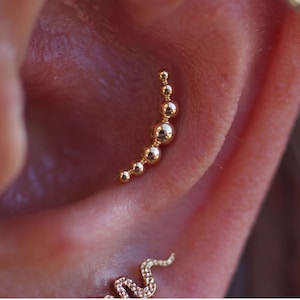 May include: A gold ear piercing jewellery piece featuring a curved row of six polished spheres that graduate in size from small to large and back to small. The metal has a reflective finish and is positioned vertically within the ear cartilage.