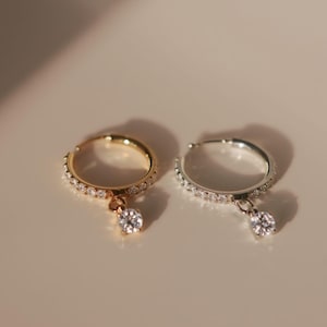 Puede incluir: Dos pendientes de aro, uno dorado y otro plateado, cada uno con una hilera de pequeñas piedras transparentes a lo largo del aro y un dije de piedra redonda transparente colgando de la parte inferior, formando un par de joyas a juego.