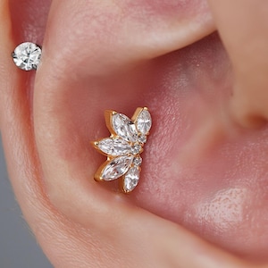 May include: Close-up of an ear with two pieces of jewellery. A small, round, clear gem is in the upper ear. A gold-coloured, floral-shaped earring with clear gems is in the lower ear.