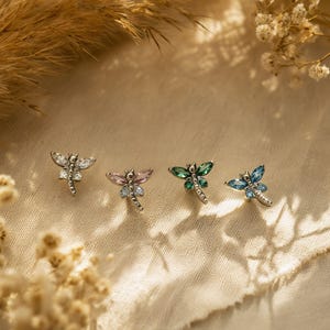 以下が含まれることがあります： 一列に並んだ4つのトンボ型スタッドピアス。各ピアスはシルバー調の金属製の本体で、羽と尾には透明、ライトピンク、エメラルドグリーン、ブライトブルーの小さなファセットカットの宝石がはめ込まれています。