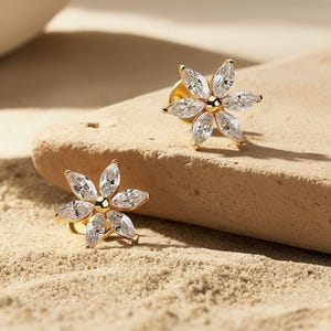 May include: A pair of gold flower stud earrings. Each earring has six clear, marquise-cut gemstones around a central gold bead. The earrings are displayed on a light brown, textured surface, with a sandy background.