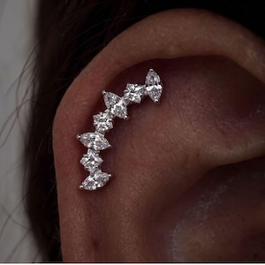 Peut inclure: Une boucle d'oreille grimpante argentée composée d'une rangée incurvée de pierres transparentes alternant entre des tailles marquise et princesse. Le bijou suit la courbe naturelle du cartilage de l'oreille avec une finition métallique brillante.