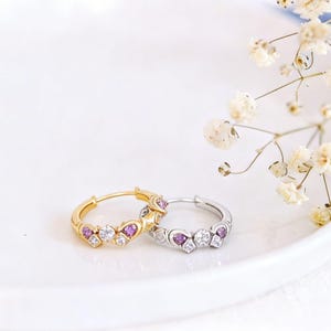 Op de afbeelding: Twee oorringen, één goudkleurig en één zilverkleurig, elk versierd met hartvormige paarse en witte edelstenen. De oorbellen liggen op een wit oppervlak, met delicate witte bloemen op de achtergrond.