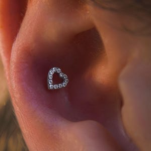 May include: A silver heart-shaped earring with clear, sparkling stones. The earring is set in a lobe piercing. The image is a close-up of the ear and earring.