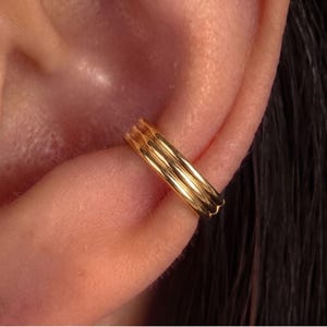 May include: A gold-toned ear cuff featuring a triple-band design with a polished, reflective finish. The jewelry piece is positioned securely along the outer edge of the ear cartilage, showcasing a smooth, rounded profile and a metallic luster.