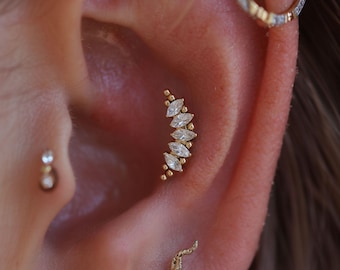 May include: A gold ear cartilage piercing featuring a curved row of five marquise-cut clear stones accented by small gold beads. The jewellery is positioned in the conch area of the ear, accompanied by a small stud and a partial hoop earring.