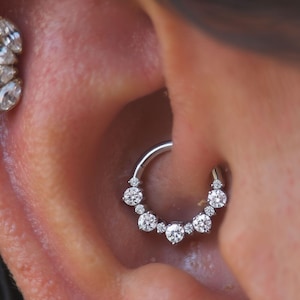 Op de afbeelding: Close-up van een oor versierd met twee sieraden. De ene is een zilveren septumring met zeven ronde, heldere edelstenen. De andere is een cluster van vijf marquise-geslepen, heldere edelstenen in een gouden zetting.