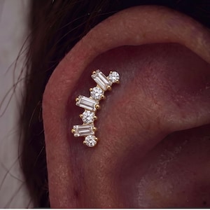 Könnte beinhalten: Ein goldener Ohrstecker für den Knorpelbereich mit einer geschwungenen Anordnung aus abwechselnd im Baguette- und Rundschliff facettierten klaren Steinen. Das Schmuckstück ist in eine Basis aus Gelbgold gefasst und folgt der natürlichen Ohrform.
