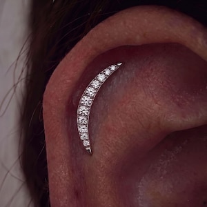 May include: A silver-toned crescent moon-shaped ear piercing jewelry piece featuring a curved row of seven small, clear, round-cut stones set along the front surface, positioned vertically along the upper cartilage of the ear.
