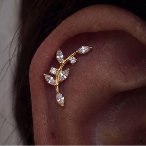 May include: A gold-toned ear cartilage piercing jewellery piece shaped like a curved branch. It features multiple marquise-cut clear stones arranged as leaves and two small round-cut clear stones set along the central gold metal stem.