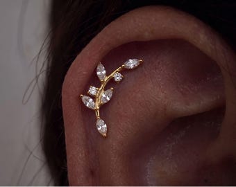 Può includere: Un gioiello per piercing alla cartilagine dell'orecchio color oro a forma di ramo curvo. Presenta diverse pietre chiare taglio marquise disposte come foglie e due piccole pietre chiare taglio rotondo lungo lo stelo centrale in metallo oro.