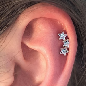 Könnte beinhalten: Nahaufnahme eines Ohrs mit drei sternförmigen Ohrringen. Jeder Ohrring hat einen klaren, facettierten Stein, der in einem silberfarbenen Metall gefasst ist. Die Ohrringe sind vertikal am Ohrknorpel angeordnet.