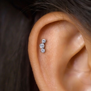 Puede incluir: Primer plano de una oreja con tres pequeñas piedras preciosas redondas y transparentes engastadas en una línea vertical como piercing. Las piedras están engastadas en un metal plateado o blanco. La oreja es de tono de piel claro.