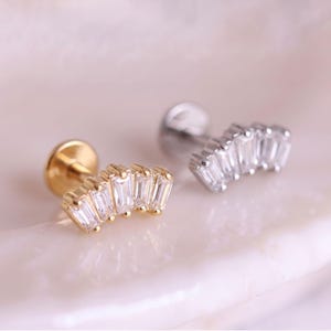 Puede incluir: Dos pendientes de botón con parte posterior plana y diseño de abanico curvo engastados con cinco piedras transparentes de corte baguette. Un pendiente es de metal dorado y el otro de metal plateado, ambos con un acabado pulido y brillante.