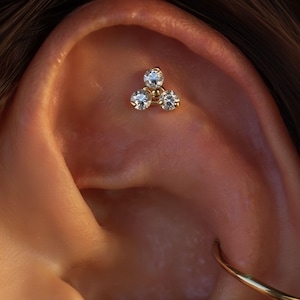 Puede incluir: Primer plano de un piercing dorado con tres gemas redondas y transparentes. El piercing está dispuesto en forma triangular. La oreja es de color piel cálido y se ve un pendiente de aro dorado.