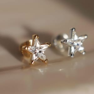 May include: Close-up of two star-shaped stud earrings. One is gold-toned with a clear, faceted center, and the other is silver-toned with a similar clear, faceted center. The earrings are on a light-colored surface.