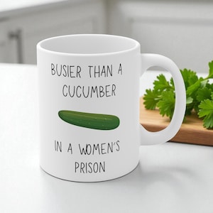 Könnte beinhalten: Weißer Keramikbecher mit Henkel, mit dem schwarzen Text "BUSIER THAN A CUCUMBER IN A WOMEN'S PRISON". Eine grüne Gurkenillustration befindet sich unter dem Text. Der Becher steht auf einer weißen Oberfläche.