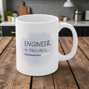 May include: White ceramic mug with a large handle. The mug features the text "ENGINEER IN PROGRESS..." in black, with a blue progress bar graphic on a light blue watercolor-style background. The mug is on a wooden surface.