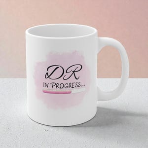 May include: White ceramic mug with a curved handle. The mug features the text "DR IN PROGRESS..." in black script, with a pink progress bar graphic on a pink watercolour-style background.