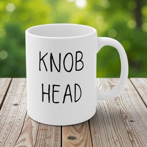 Könnte beinhalten: Weiße Keramik-Tasse mit den schwarzen Worten "KNOB HEAD" mittig auf der Vorderseite. Die Tasse hat einen gebogenen Henkel und steht auf einer Holzoberfläche.