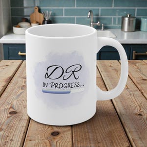 May include: White ceramic mug featuring the text "DR IN PROGRESS..." in black script and a blue progress bar design. The mug has a curved handle. The background is a kitchen setting.
