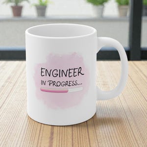 Peut inclure: Mug en céramique blanche avec une anse incurvée. Le mug présente le texte "ENGINEER IN PROGRESS..." en noir, avec un graphique de barre de chargement rose sur un fond rose de style aquarelle.
