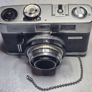 May include: A vintage silver and black camera with a lens and a chain. The camera has the text "electric-CR" and "signette" on it. The camera is on a light grey surface.