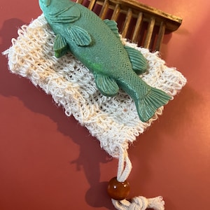 May include: A green fish-shaped soap bar rests on a natural fiber soap saver. The soap saver is a light beige color and has a drawstring closure with a wooden bead. A small wooden rack is in the background.