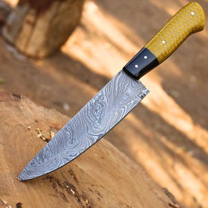 May include: A Damascus steel chef's knife with a patterned blade and a yellow honeycomb-patterned handle. The knife has a black bolster and is set against a wooden surface, showcasing its craftsmanship.