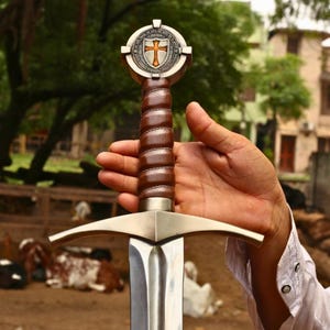 May include: A medieval sword with a brown leather-wrapped handle and a silver blade. The hilt has a decorative emblem with a cross design. The sword is held against a blurred outdoor backdrop.