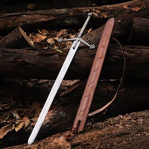 Può includere: Una spada d'argento e acciaio con un fodero in pelle. La spada ha un'elsa e una guardia dettagliate. Il fodero è marrone con cuciture decorative e una tracolla. La spada e il fodero poggiano su un letto di tronchi e foglie.