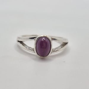 May include: A silver ring with a large, oval, deep purple gemstone. The ring's band splits into two parallel lines on either side of the gemstone, creating an open design. The gemstone is the focal point, set in a simple, elegant style.