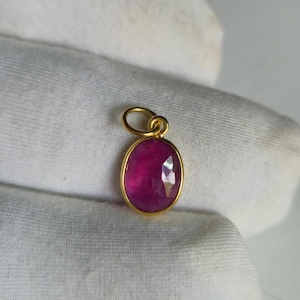 May include: A gold pendant featuring a faceted, oval-shaped, deep pink gemstone. The gemstone is set in a gold bezel and has a small loop at the top for a chain. The pendant is displayed on white fabric.