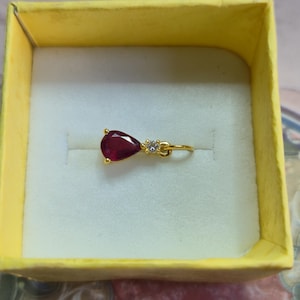 Puede incluir: Un colgante de oro con una piedra preciosa roja oscura en forma de lágrima y dos pequeñas piedras preciosas claras. El colgante está en una caja amarilla con interior blanco.