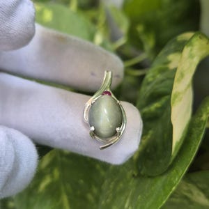 Op de afbeelding: Een zilveren hanger met een gladde, ovaalvormige grijze kattenoog-edelsteen in een zetting met pootjes. Een kleine, ronde, donkerroze edelsteen is aan de bovenkant geplaatst, geïntegreerd in het gebogen, gepolijste zilveren metalen frame.
