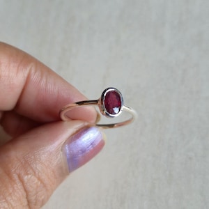 Può includere: Un anello in argento con una singola pietra preziosa di rubino rosso intenso di forma ovale, montata in una lunetta liscia. La fascia è sottile e lucida, caratterizzata da un design essenziale che mette in risalto la superficie sfaccettata.