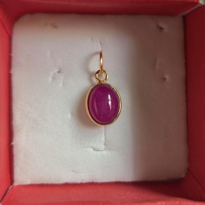 May include: A gold pendant featuring an oval-shaped, smooth, deep pink cabochon gemstone set in a simple gold bezel. The pendant includes a small circular gold jump ring at the top for attachment to a chain, presented in a box.