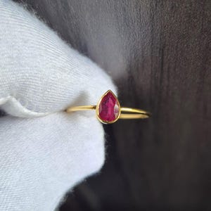 Könnte beinhalten: Ein Goldring mit einem einzelnen birnenförmigen, tiefrosa Edelstein in einer glatten Zargenfassung. Die Ringschiene ist dünn und poliert und unterstreicht mit ihrem schlichten Design die leuchtende Farbe und Tropfenform des Steins.