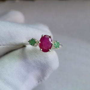 May include: A silver ring featuring a large oval-cut ruby gemstone as the centerpiece, flanked by two smaller emeralds. The ruby is a deep red color, and the emeralds are a vibrant green. The ring is set on a white background.