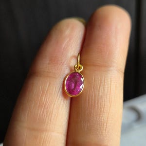 May include: A small, oval ruby pendant in a gold setting. The ruby is a bright pink hue, set in a simple gold frame. The pendant is designed to be worn as a necklace charm. The pendant's dimensions are approximately 1 cm.