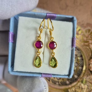 May include: A pair of gold-tone drop earrings featuring a round, faceted magenta stone at the top and a pear-shaped, faceted lime green stone at the bottom. The earrings are suspended from gold-tone hooks and presented in a light blue box.