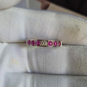 Könnte beinhalten: Ein silberner Ring mit einer Reihe leuchtend roter Edelsteine, die sich mit klaren, funkelnden Steinen abwechseln. Der Ring ist vor einem weichen, weißen Stoff platziert, der den Kontrast zwischen den hellen Edelsteinen und dem kühlen Metall hervorhebt.