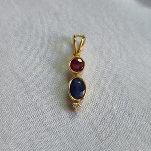 Könnte beinhalten: Goldanhänger mit einem Rubin und einem Saphir, sowie einem kleinen Diamantakzent. Der Rubin ist rund und der Saphir oval. Der Anhänger hängt an einer goldenen Öse.