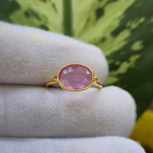 Puede incluir: Un anillo de oro con una piedra preciosa rosa de forma ovalada y tallada, montada en un engaste liso. La banda es fina y pulida, mostrando un tono de oro amarillo brillante que contrasta con el tono rosa claro translúcido de la piedra.