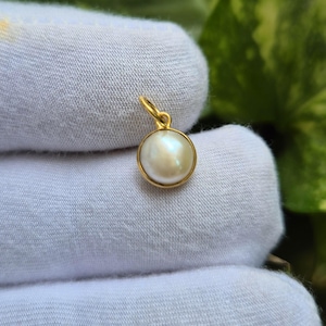 Puede incluir: Un pequeño colgante de perla blanca y redonda engastado en un bisel de metal dorado pulido. El colgante presenta un sencillo lazo circular dorado en la parte superior para su fijación, mostrando una superficie lisa y brillante en la perla.