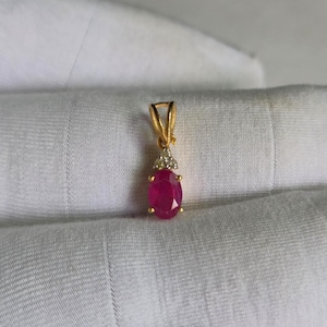 May include: A gold pendant featuring an oval-shaped, deep pinkish-red gemstone set in a four-prong mounting. Above the main stone, a small, triangular-shaped accent piece is encrusted with three tiny, clear, sparkling white stones.